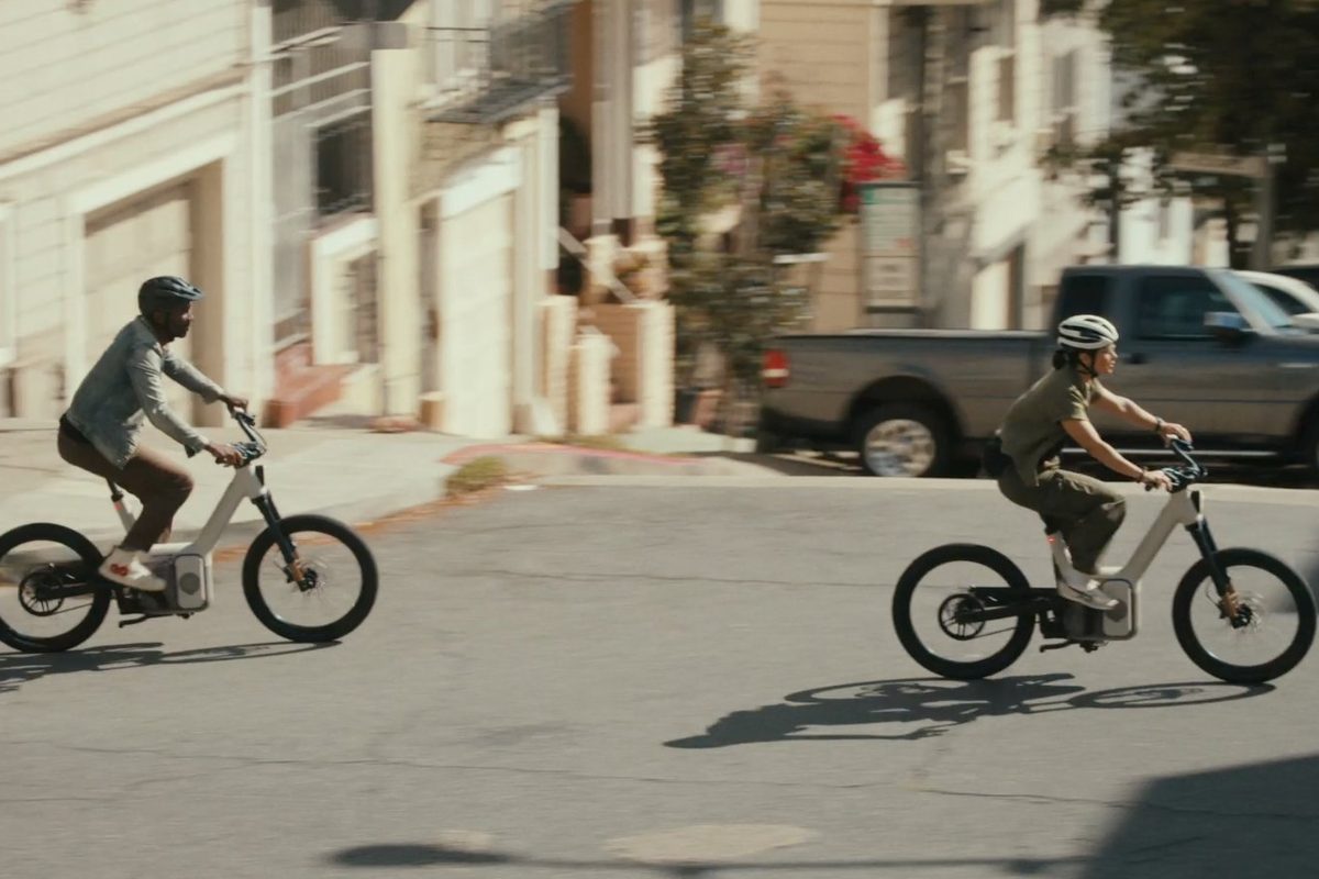 Two riders on ALSO electric bikes riding through a city street intersection
